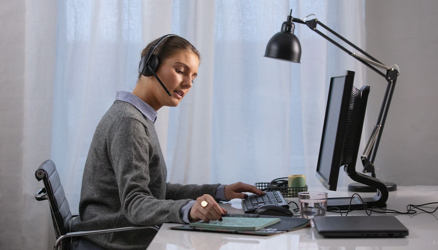 Woman using headset at desk with computer, keyboard, and lamp for remote work or customer support